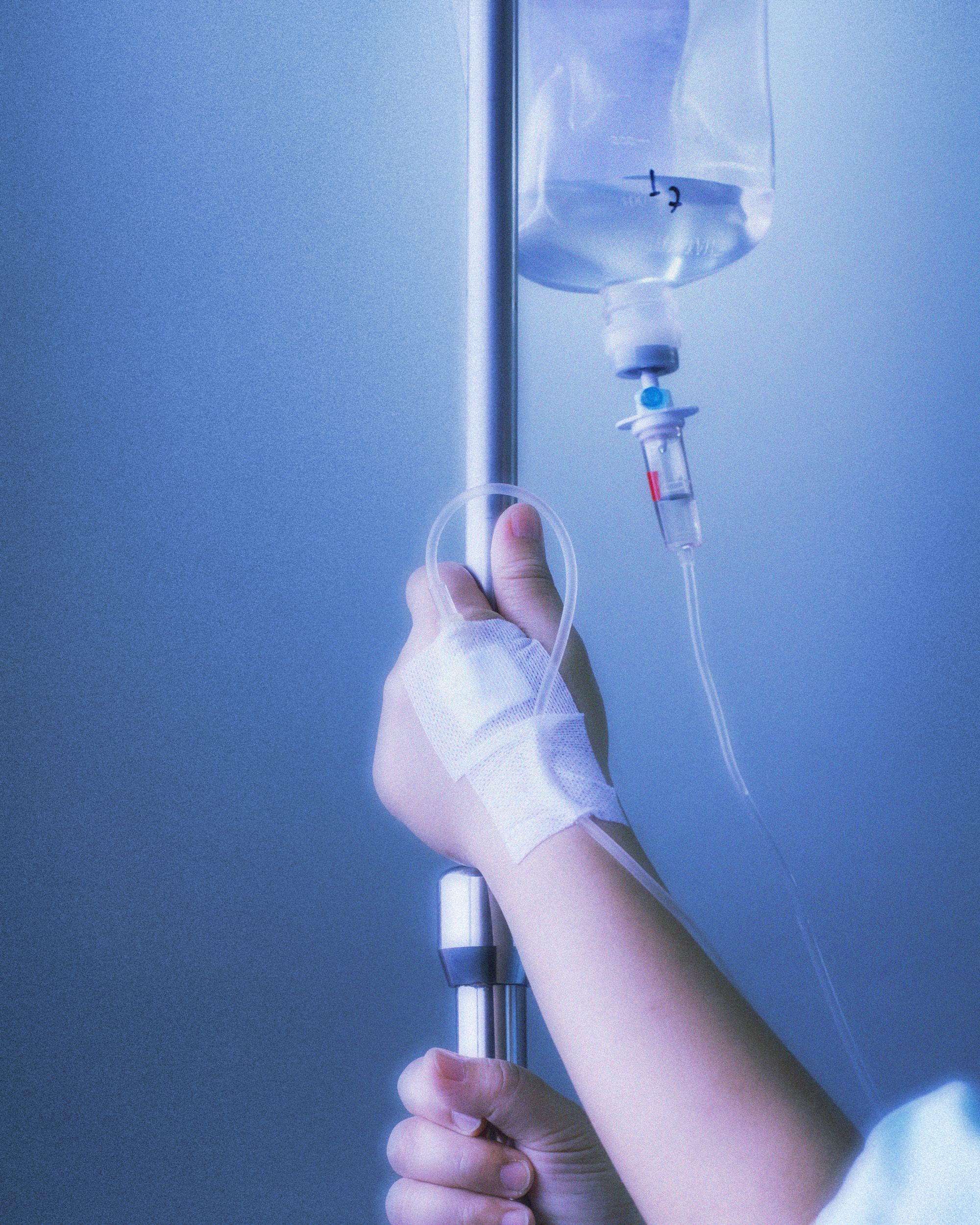 Image: An IV bag of chemo hangs while hands taped up to tubes hold the pole.